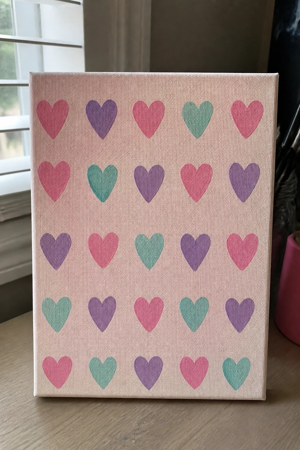 Pink canvas with grid of pink, purple, and green hearts on wooden table