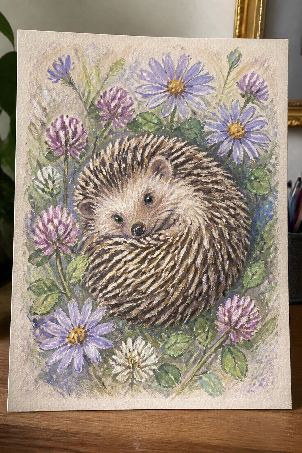 Watercolor painting of curled hedgehog amid purple daisies, pink clover, and green leaves