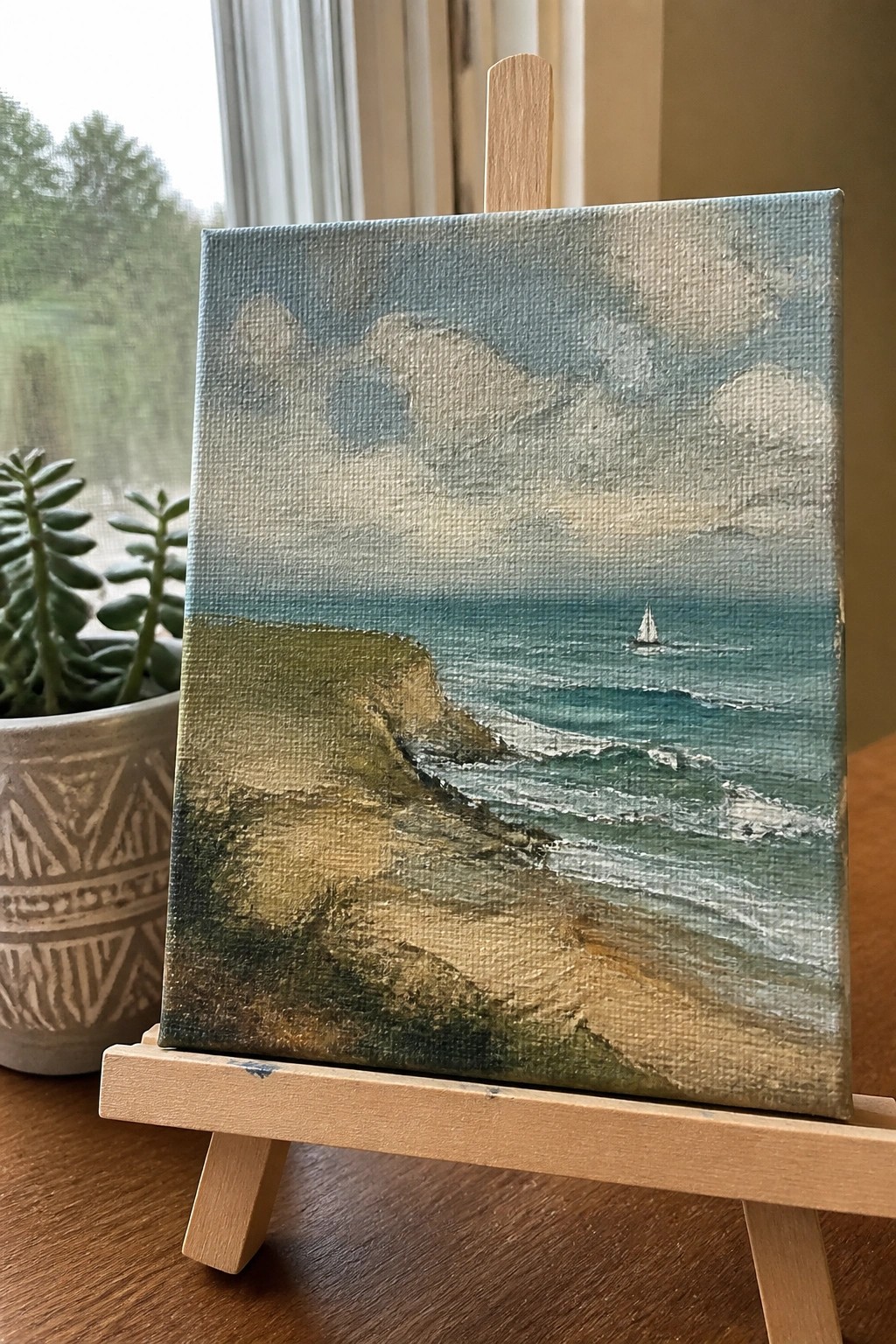 Small seascape painting on easel showing cliffs, waves, and distant sailboat beside plant.