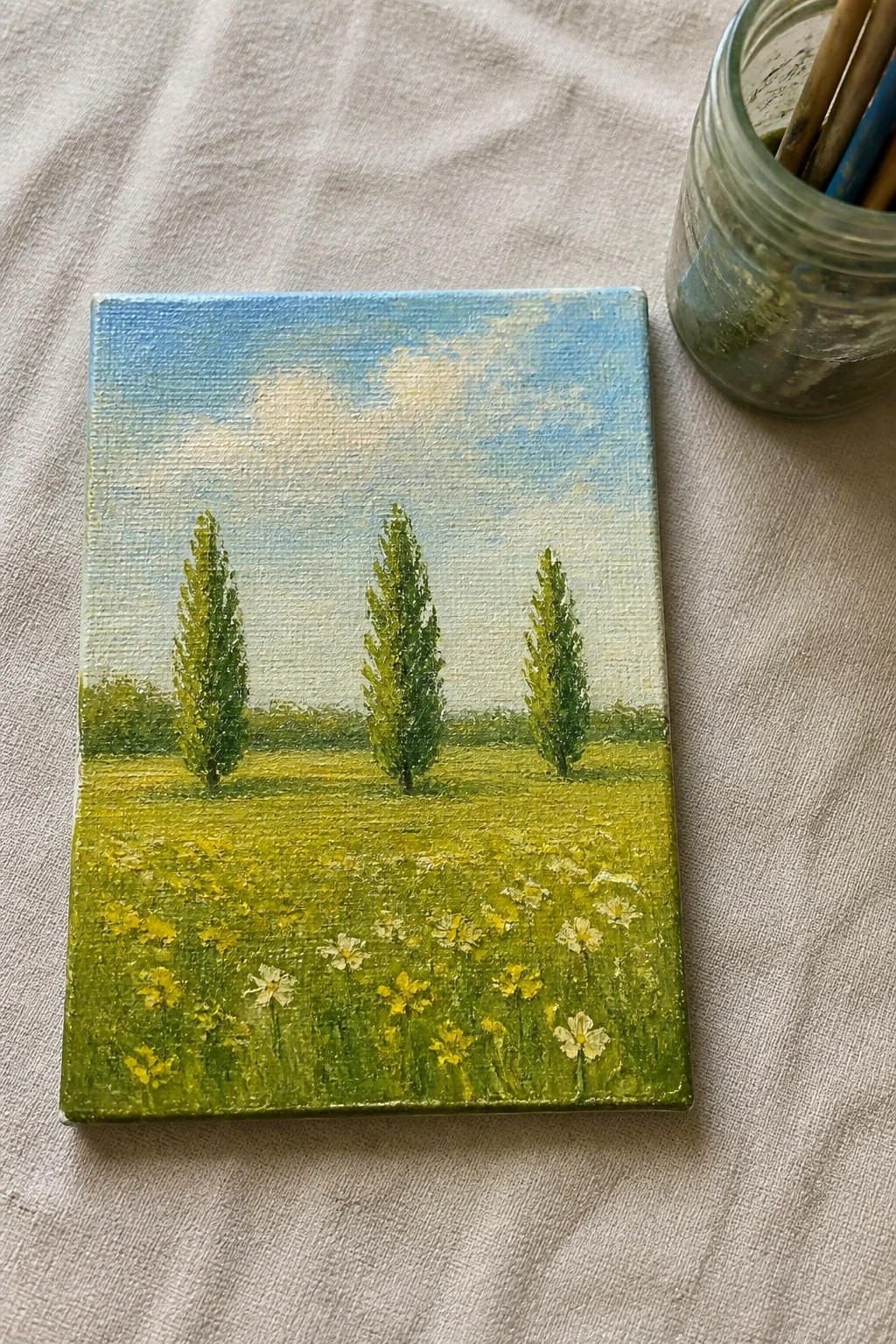 Small painting of green meadow with three trees, yellow flowers, blue sky beside brush jar.