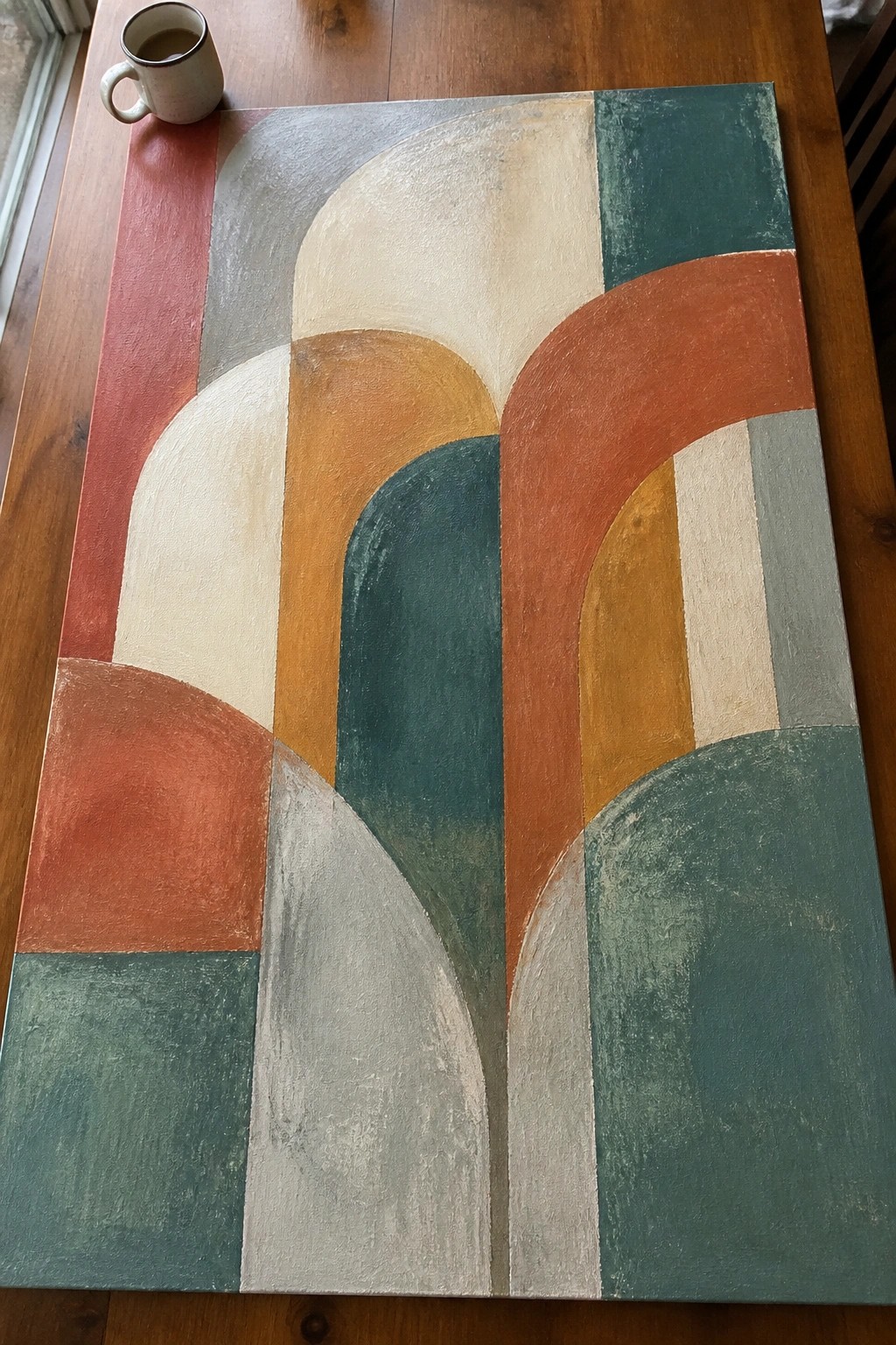 Abstract painting with curved shapes in red, teal, and orange beside coffee mug on wood table