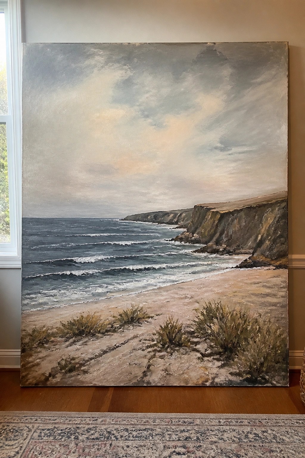 Large oil painting of waves crashing on a sandy beach beside rocky cliffs under cloudy skies.