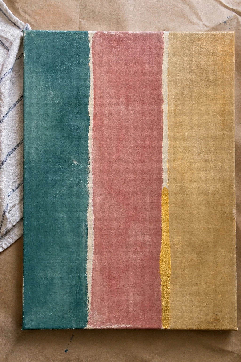 Abstract canvas with vertical teal, pink, and yellow stripes on brown paper beside striped cloth.
