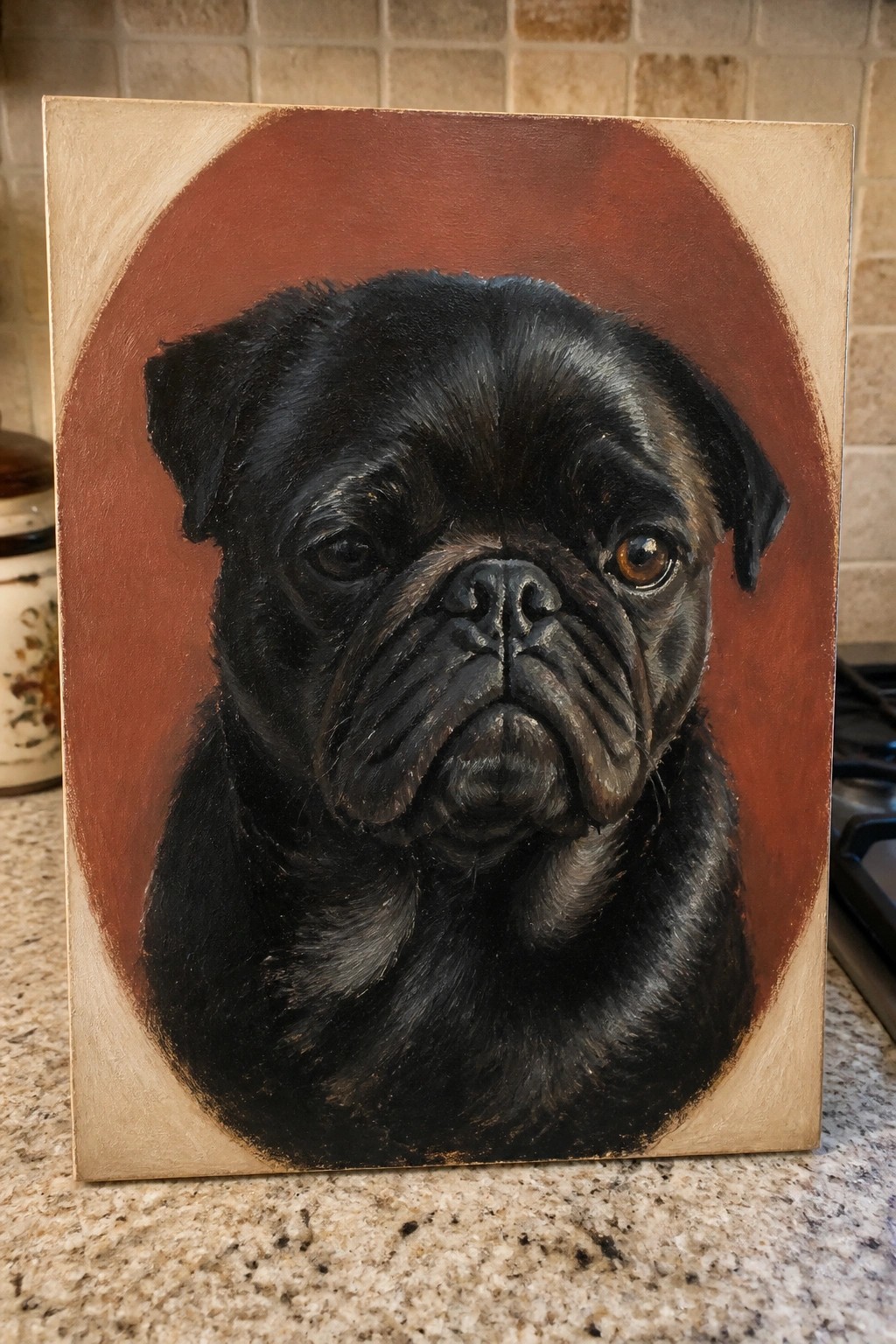 Black pug painting on canvas with reddish-brown circular background on kitchen counter.
