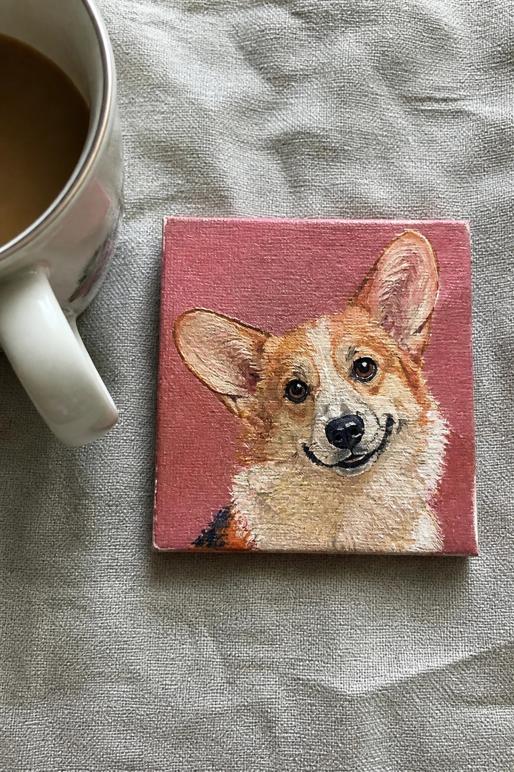 Corgi portrait painting on pink canvas beside white coffee mug on gray cloth
