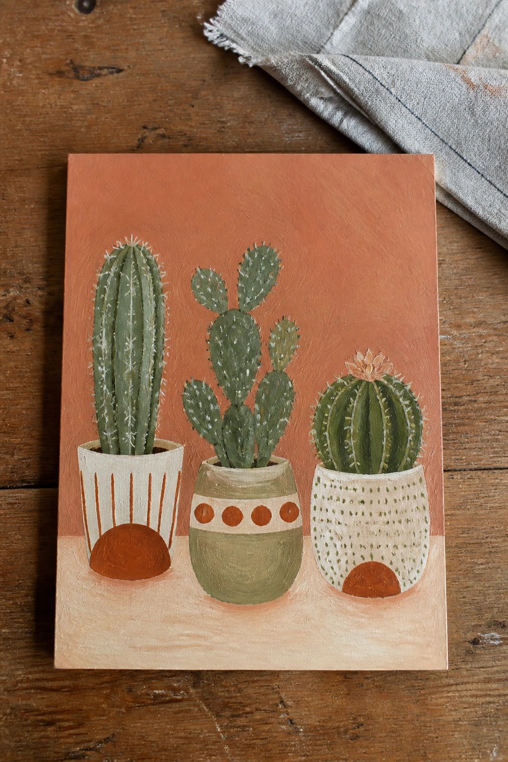 Three potted cacti in oil paint against a solid terracotta background, showing a tall columnar cactus on the left, a prickly pear in the center, and a round cactus with a flower on the right.