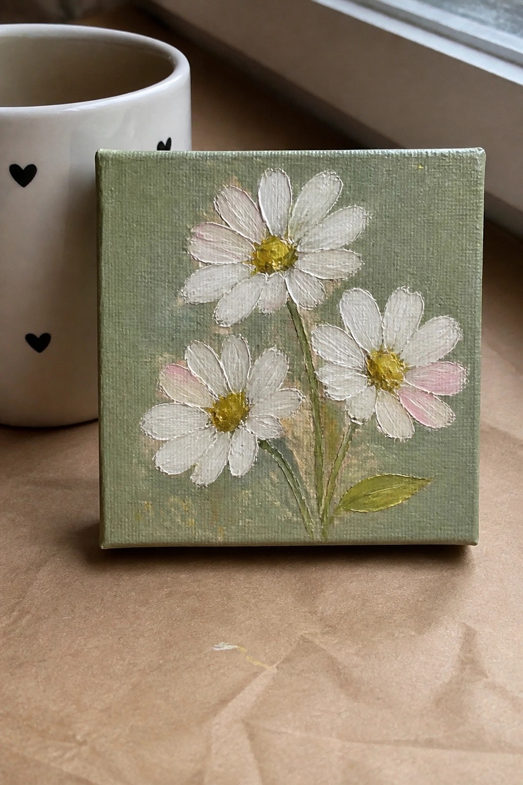 Small square canvas with three white daisies on green beside heart-patterned mug
