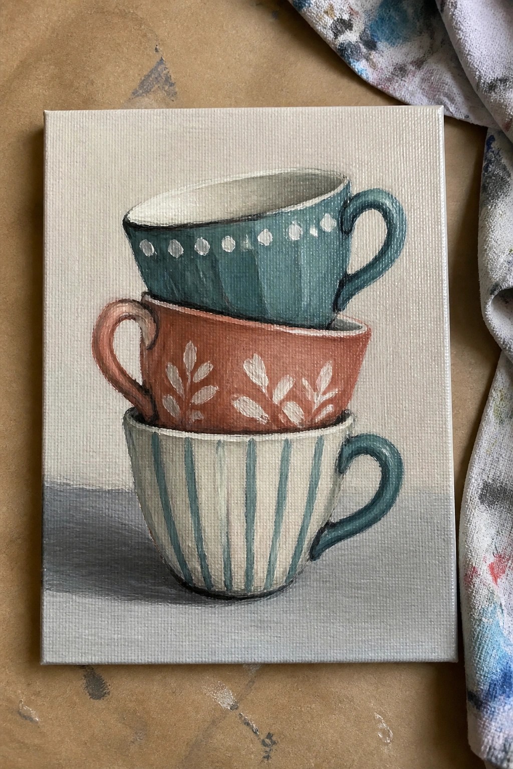 Three stacked teacups in teal, coral, and striped patterns on canvas with shadow.