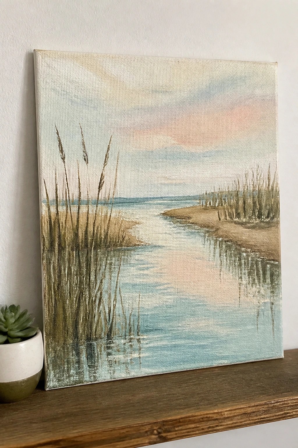 Canvas painting of marsh with reeds, calm water, and pink sky beside potted succulent on wood