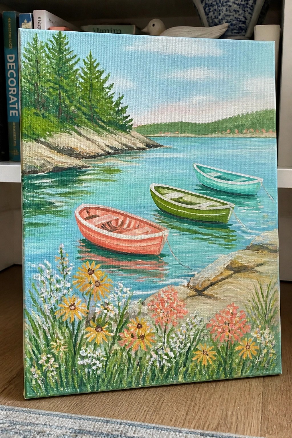 Three colorful rowboats on calm blue water by flower-lined rocky shore with pines
