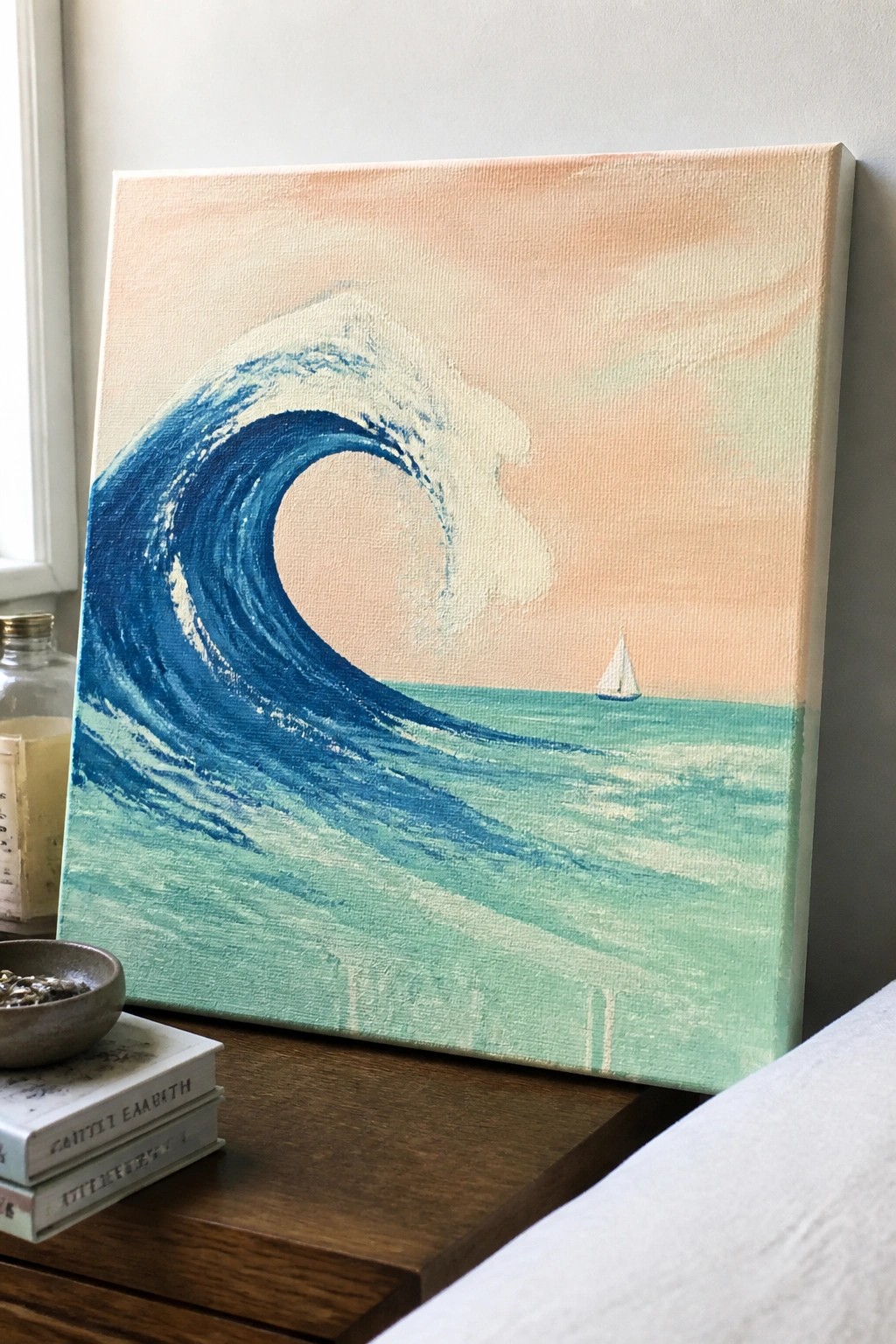 Canvas painting of curling blue wave with sailboat on peach sky beside books