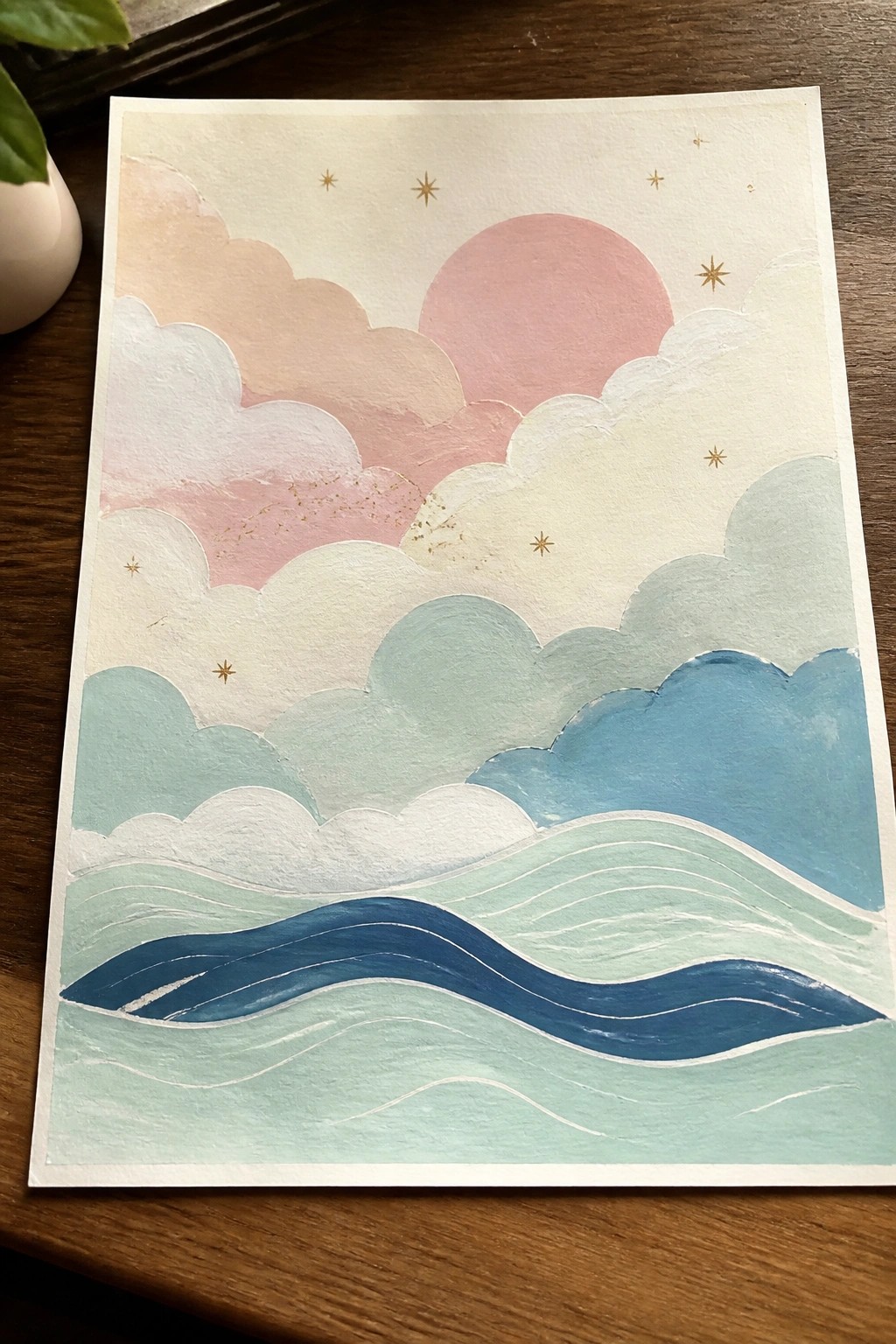A painting shows layered pastel clouds, a pink sun, gold stars, and blue-green wavy ocean on paper.