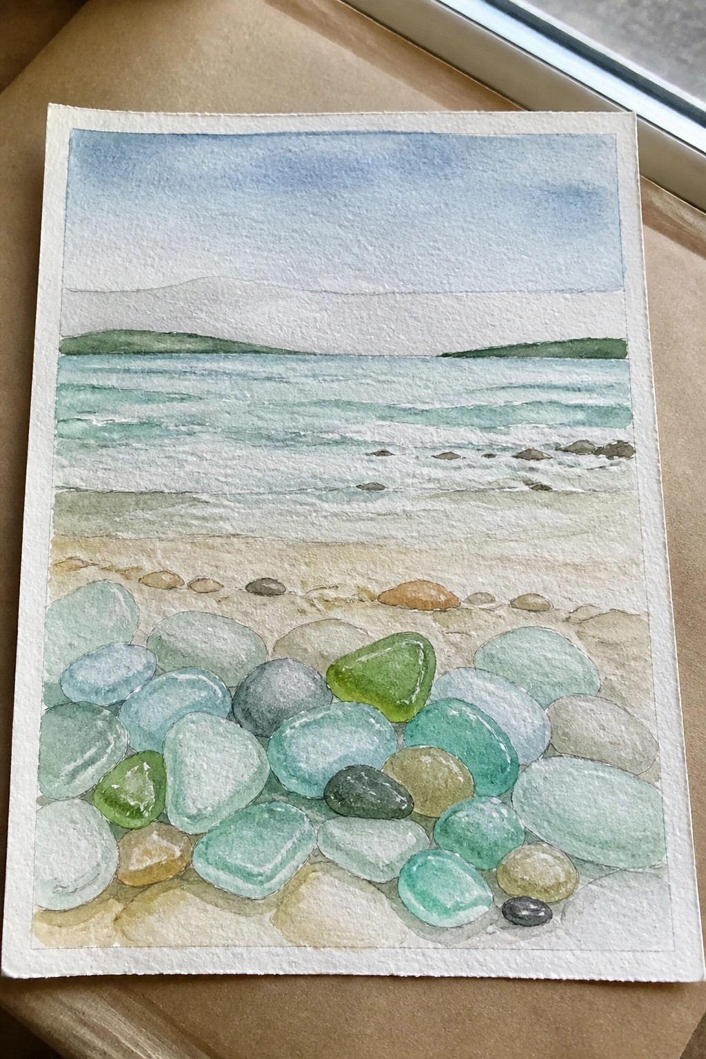 Watercolor beach scene with colorful pebbles, turquoise waves, and distant green hills.