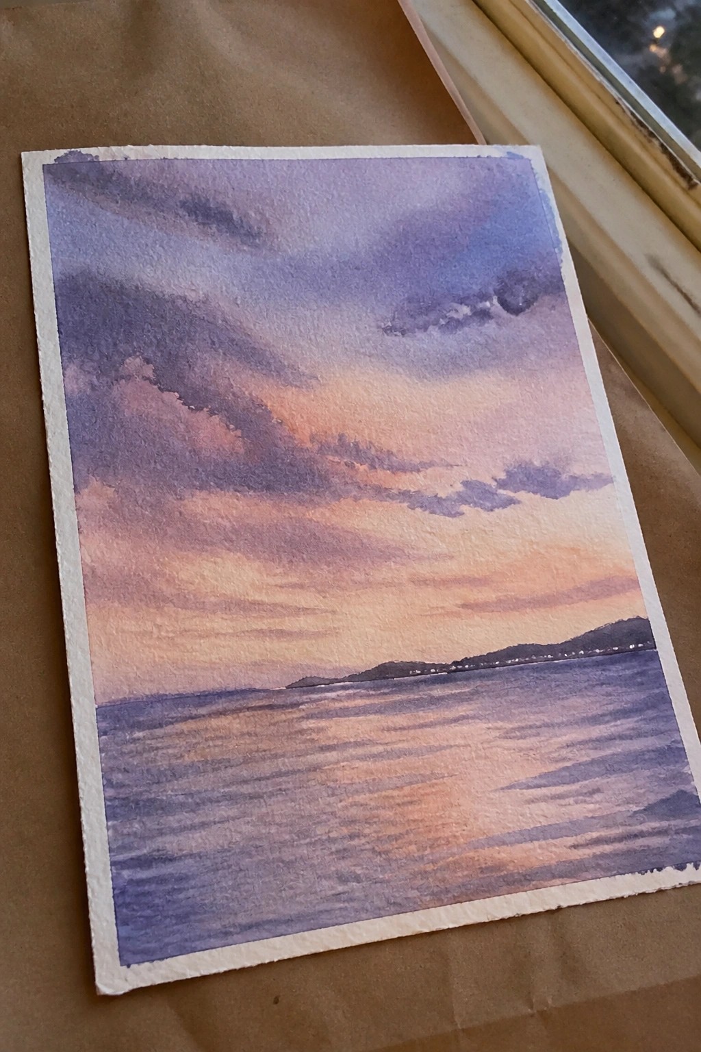 Watercolor painting of purple-orange sunset sky over rippled water with distant hills.