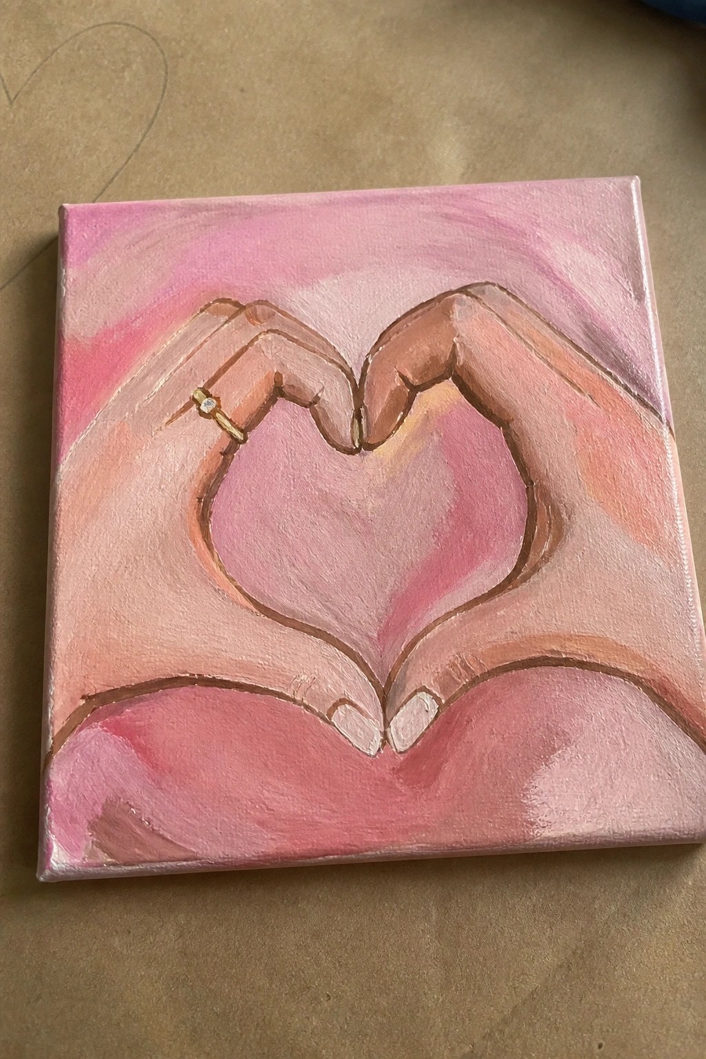 Pink acrylic painting of hands forming a heart with gold diamond ring on canvas.