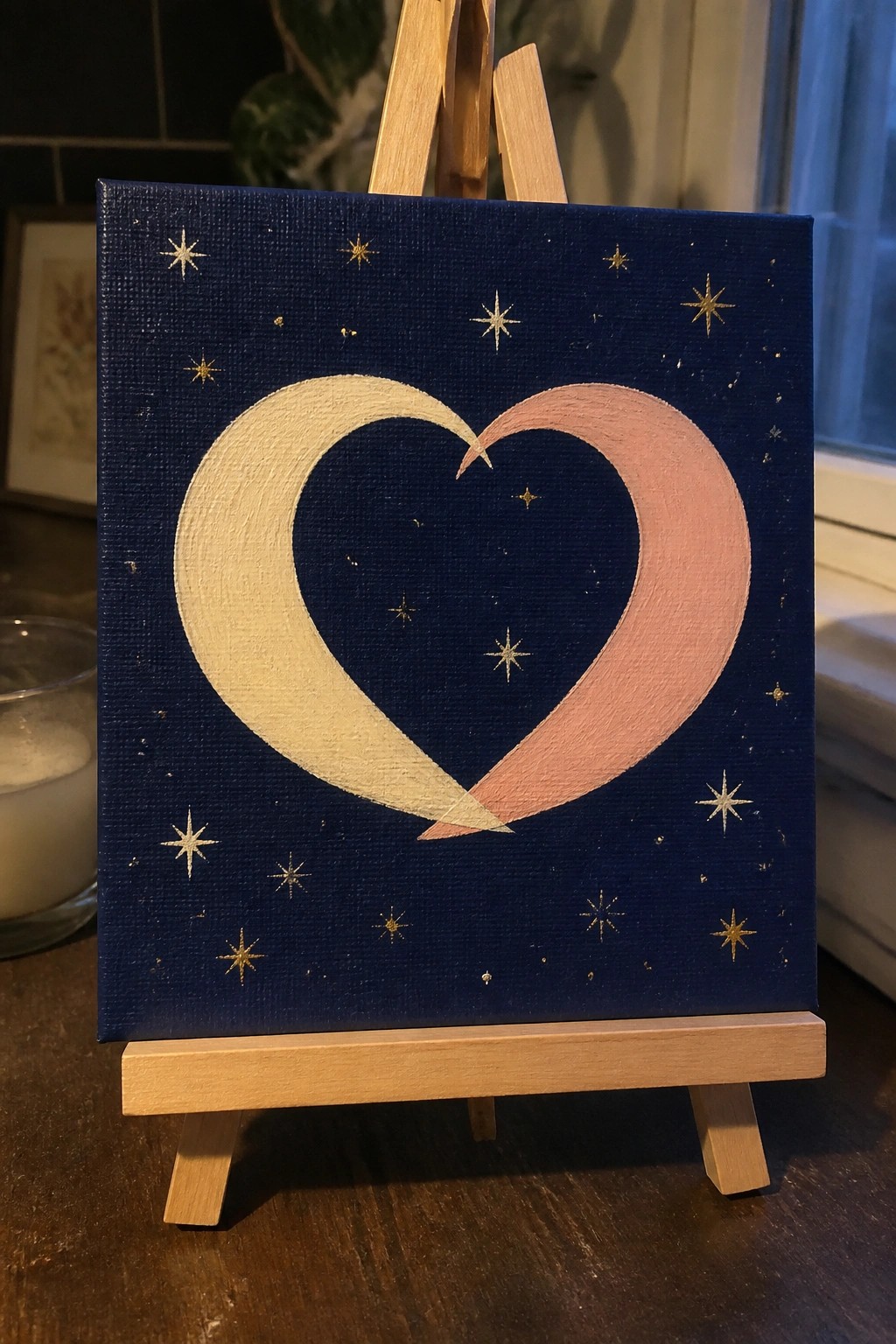 Small canvas on easel shows white and pink crescent moons forming heart with gold stars