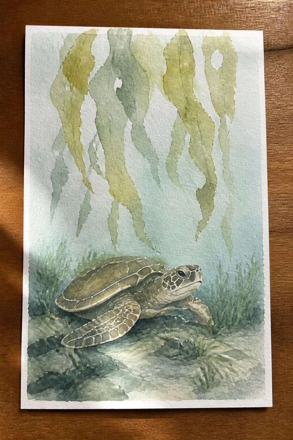 Watercolor sea turtle swimming underwater amid flowing green seaweed over rocky seabed.