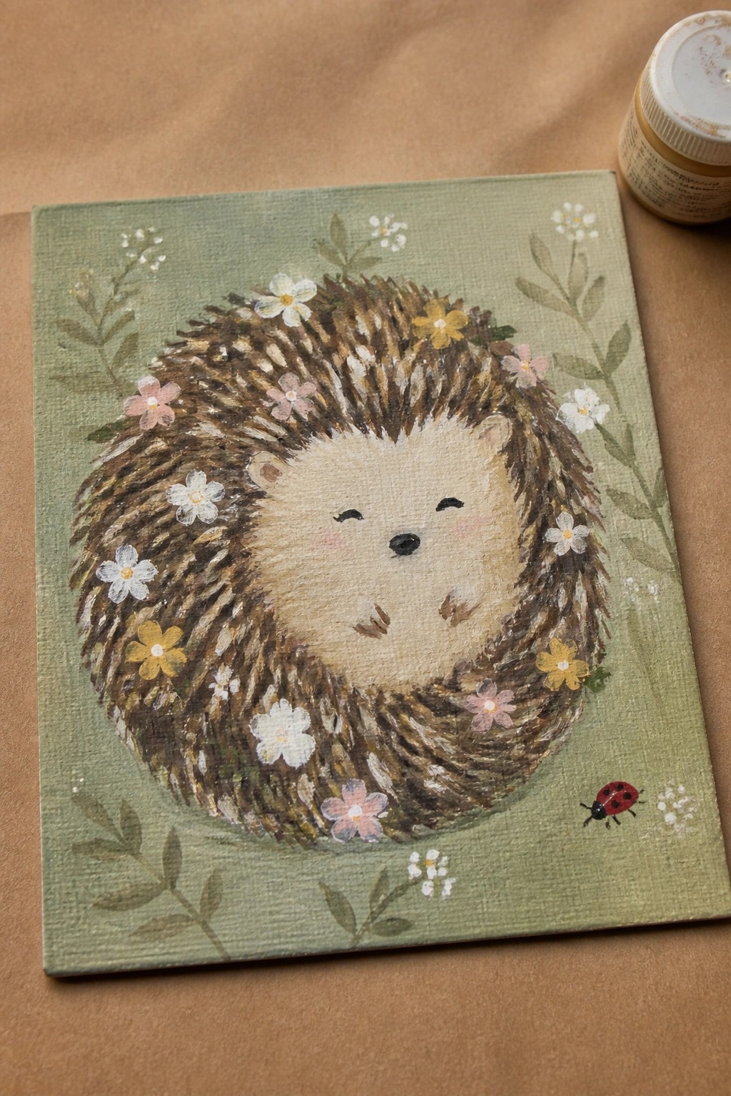 A round hedgehog painted in brown tones with white, pink, and yellow flowers on its spines sits centered on a green canvas with scattered leaves and a small ladybug.