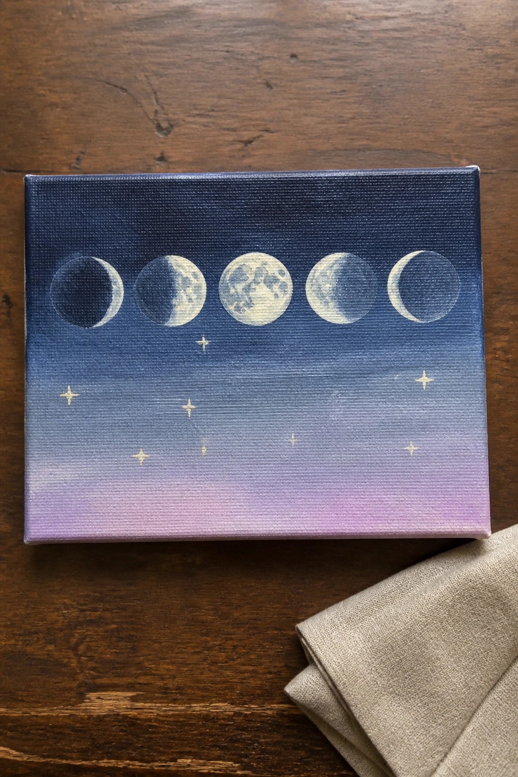 Moon phases painting on canvas with blue-purple gradient sky and gold stars on wood