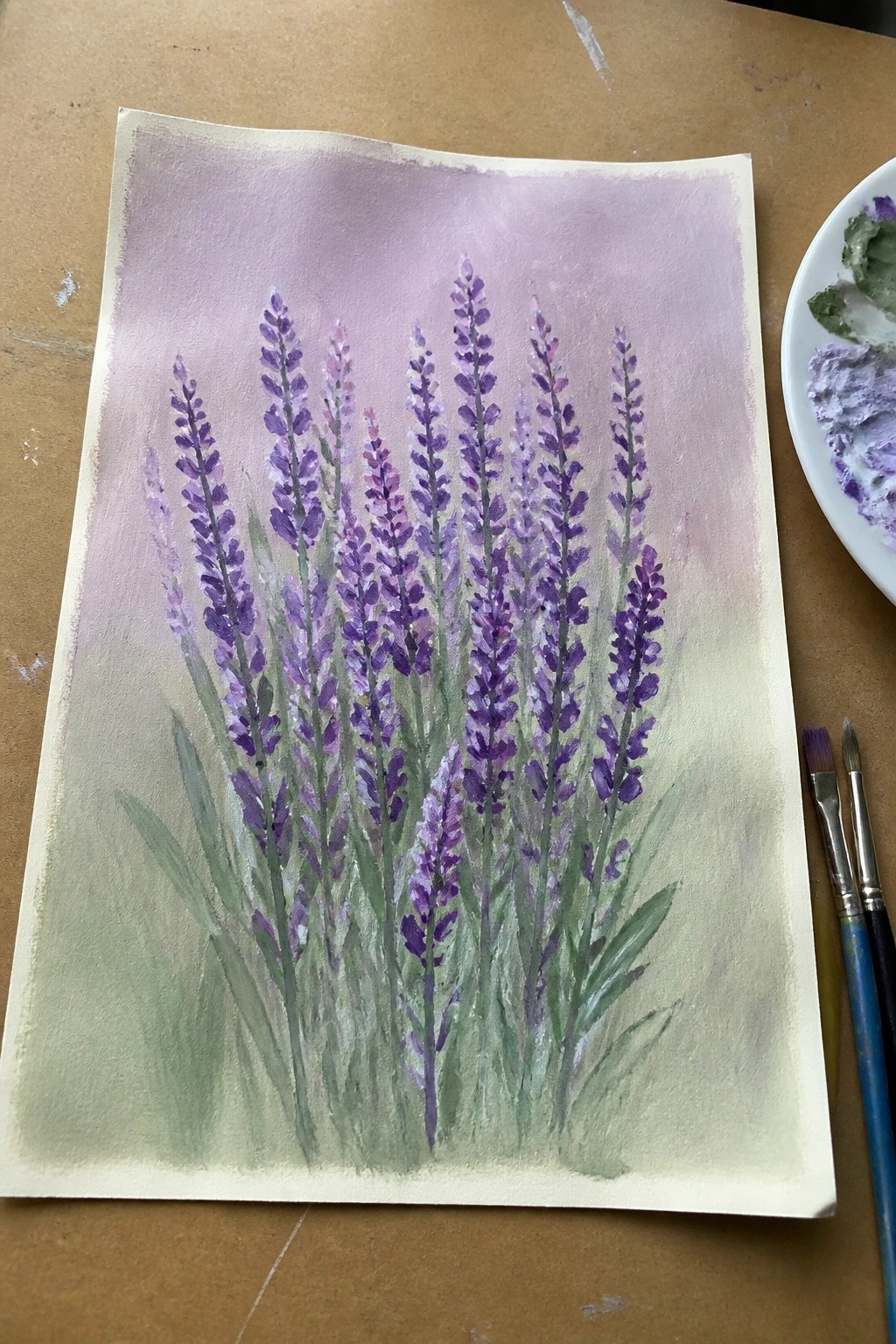 Watercolor painting of purple lavender stalks with green leaves beside paint palette and brushes