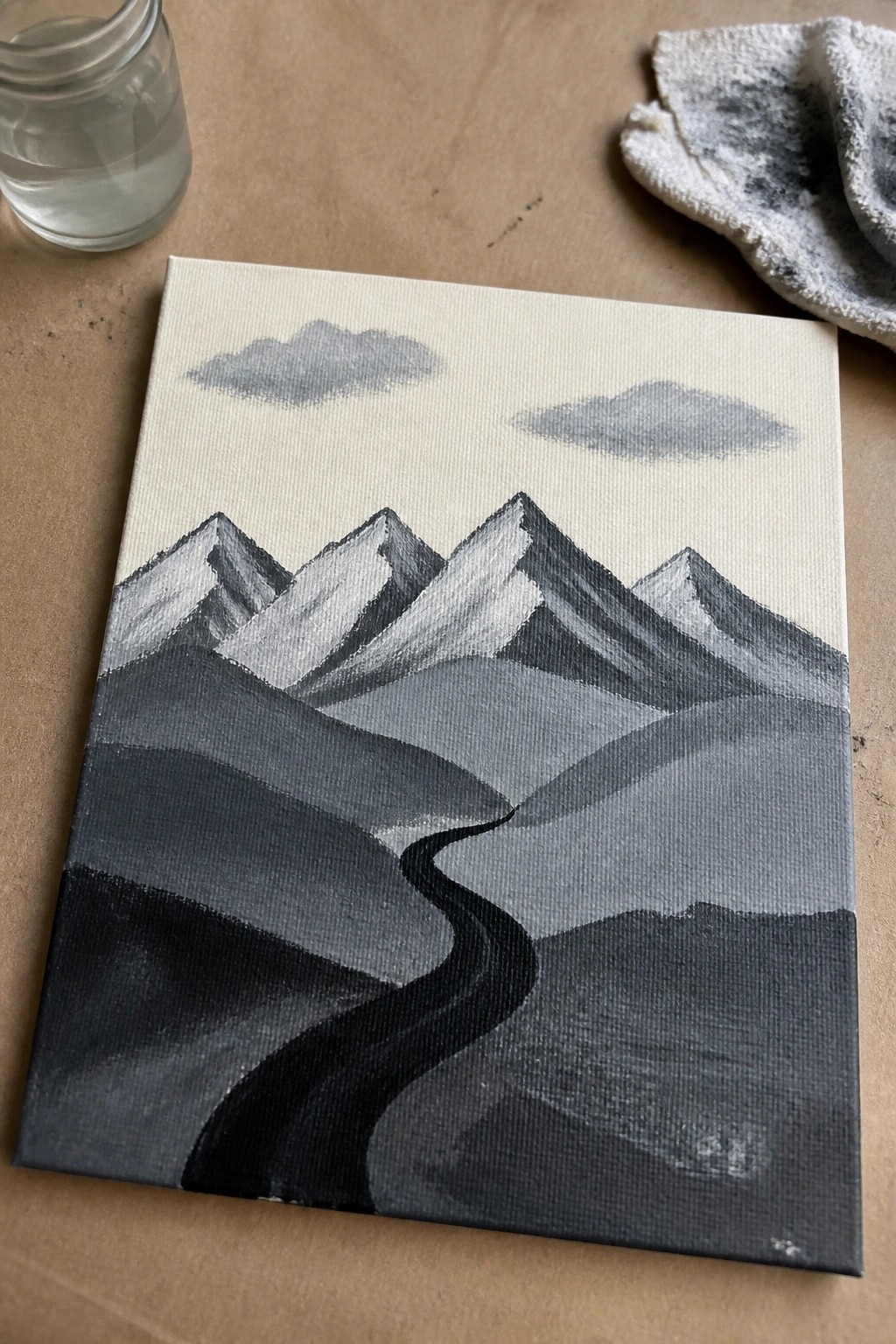 Grayscale mountain painting with winding path on canvas beside water jar and rag