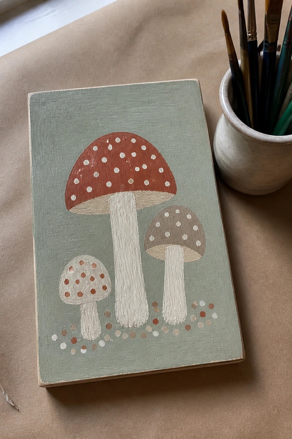 Sage green painted wood with three dotted mushrooms beside paintbrushes in a cup.