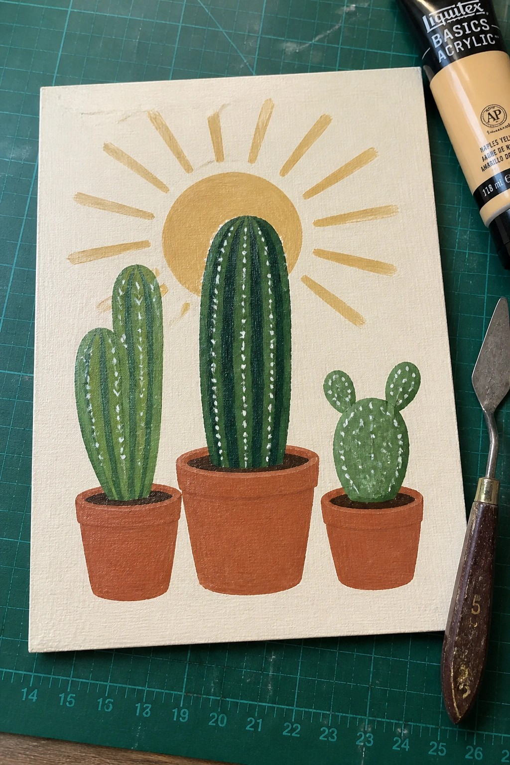 Three potted cacti on canvas with yellow sun, paint tube, and palette knife