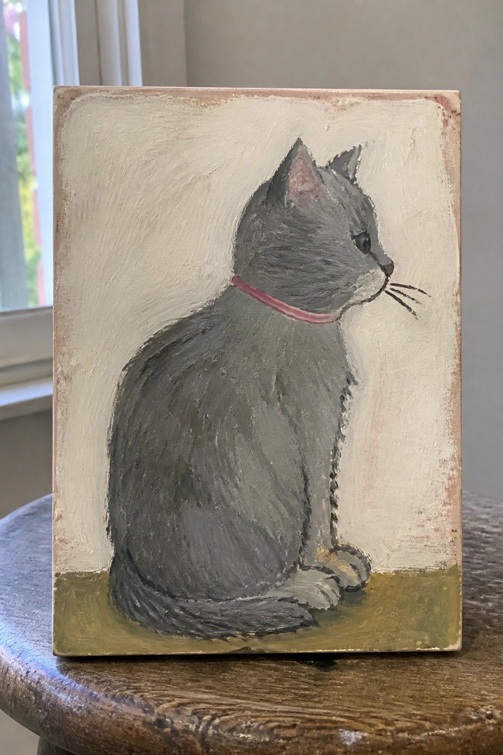 Roughly painted gray cat with pink collar sitting on white and green canvas atop wood