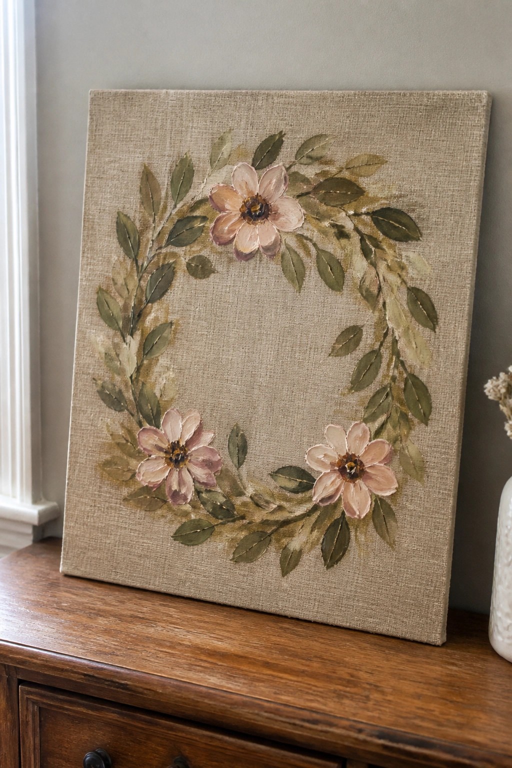 Beige canvas with pink floral wreath and green leaves on wooden dresser.