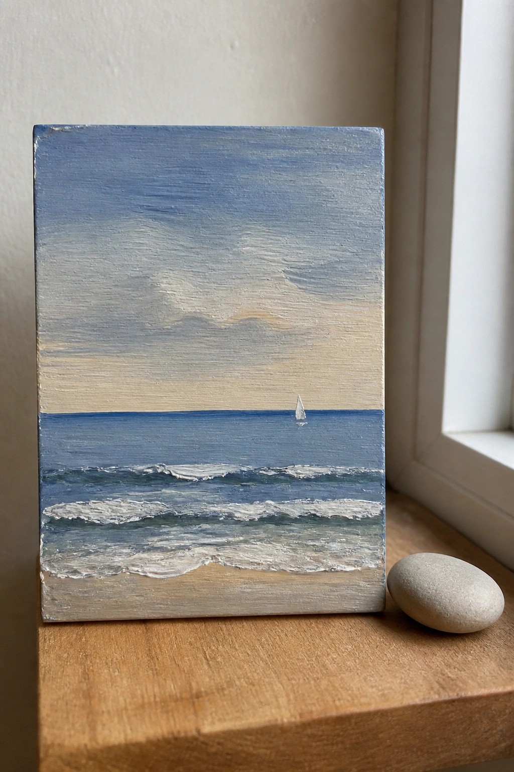 Small canvas seascape with blue sky, waves, sailboat, and beach beside a stone.