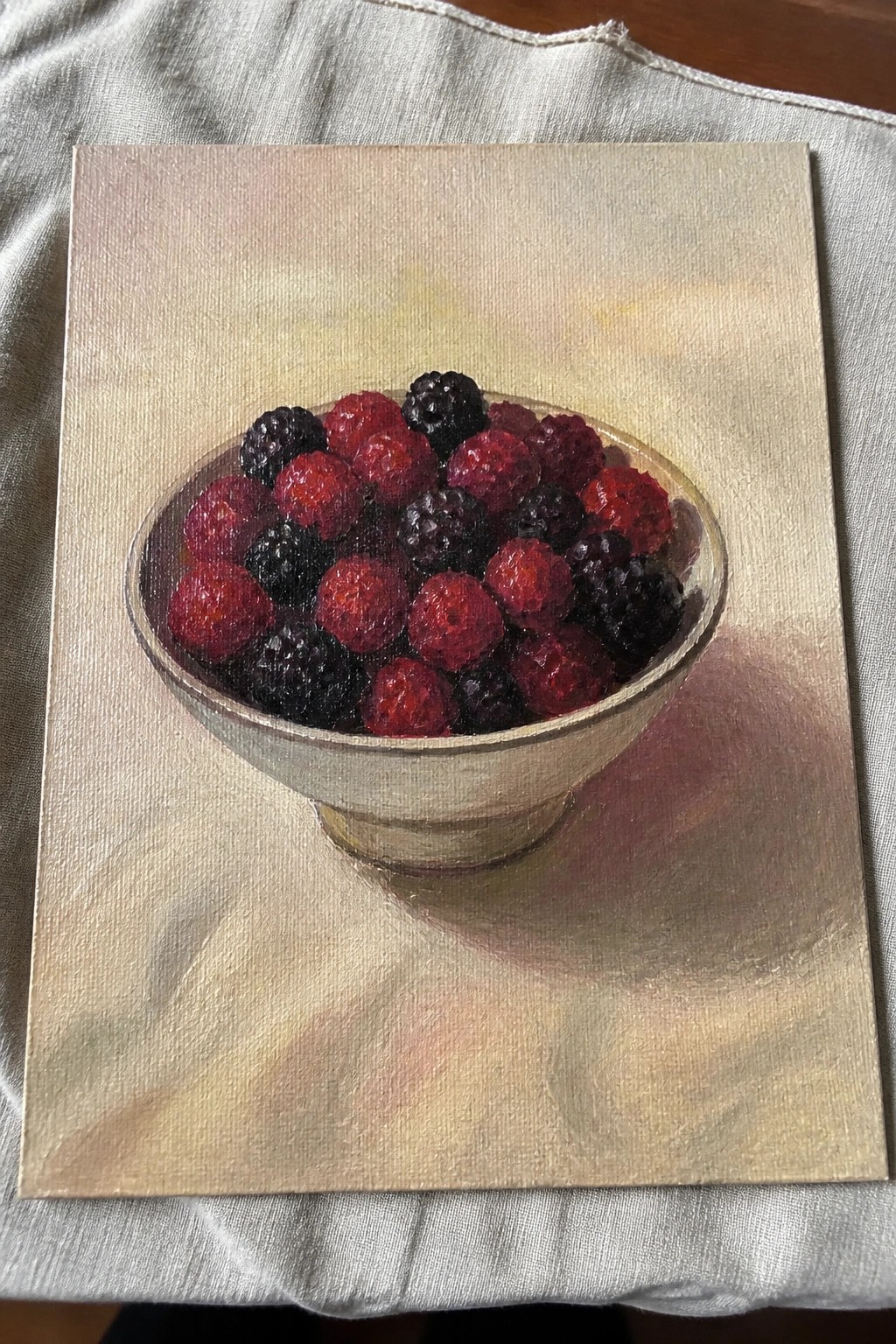 Realistic still life painting of a bowl with raspberries and blackberries on gray cloth.