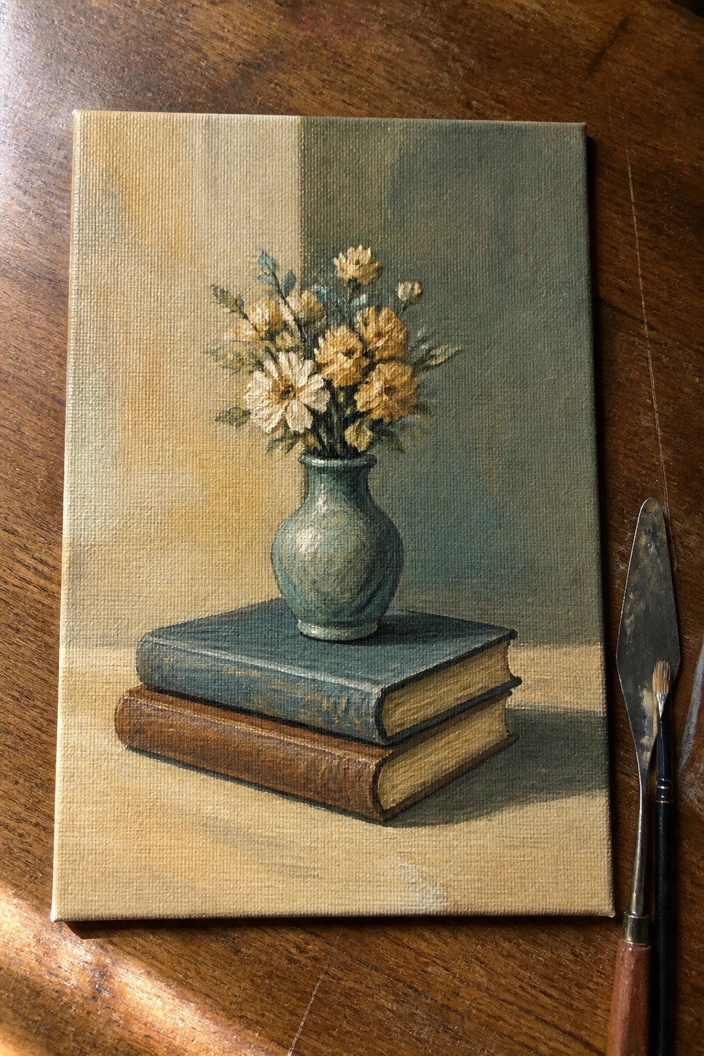 Still life painting of yellow flowers in vase atop stacked books beside palette knife