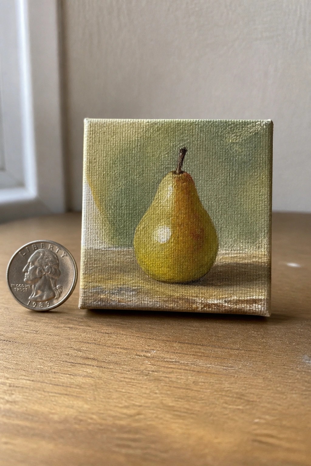 Small canvas painting of yellow pear beside 1989 quarter on wooden table