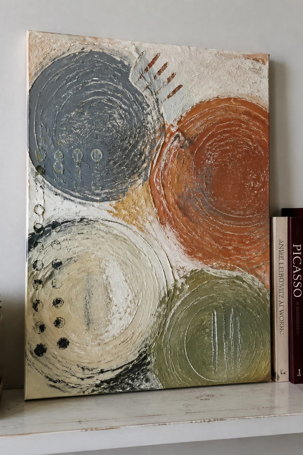 Abstract painting with textured gray, orange, beige, and green circles beside books.