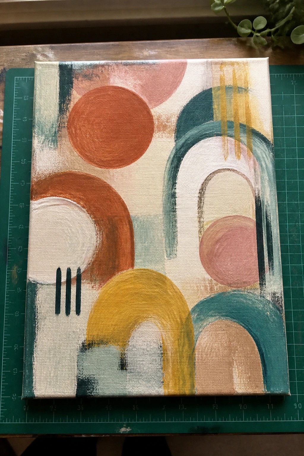 Abstract painting with orange circles, teal arches, yellow shapes on canvas atop green mat.