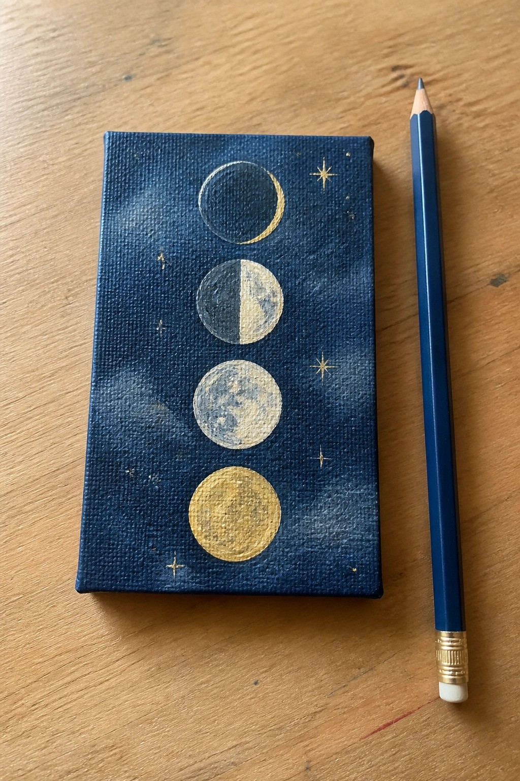 Small dark blue canvas with gold moon phases and stars beside blue pencil