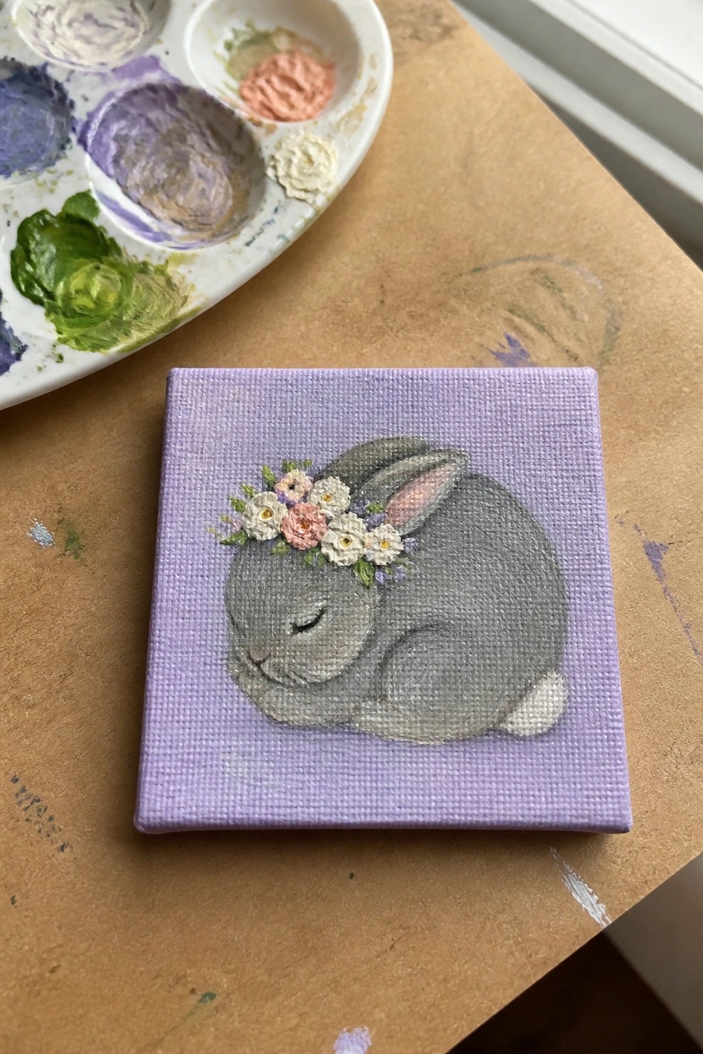 Miniature canvas shows sleeping gray rabbit with flower crown on purple background beside palette.