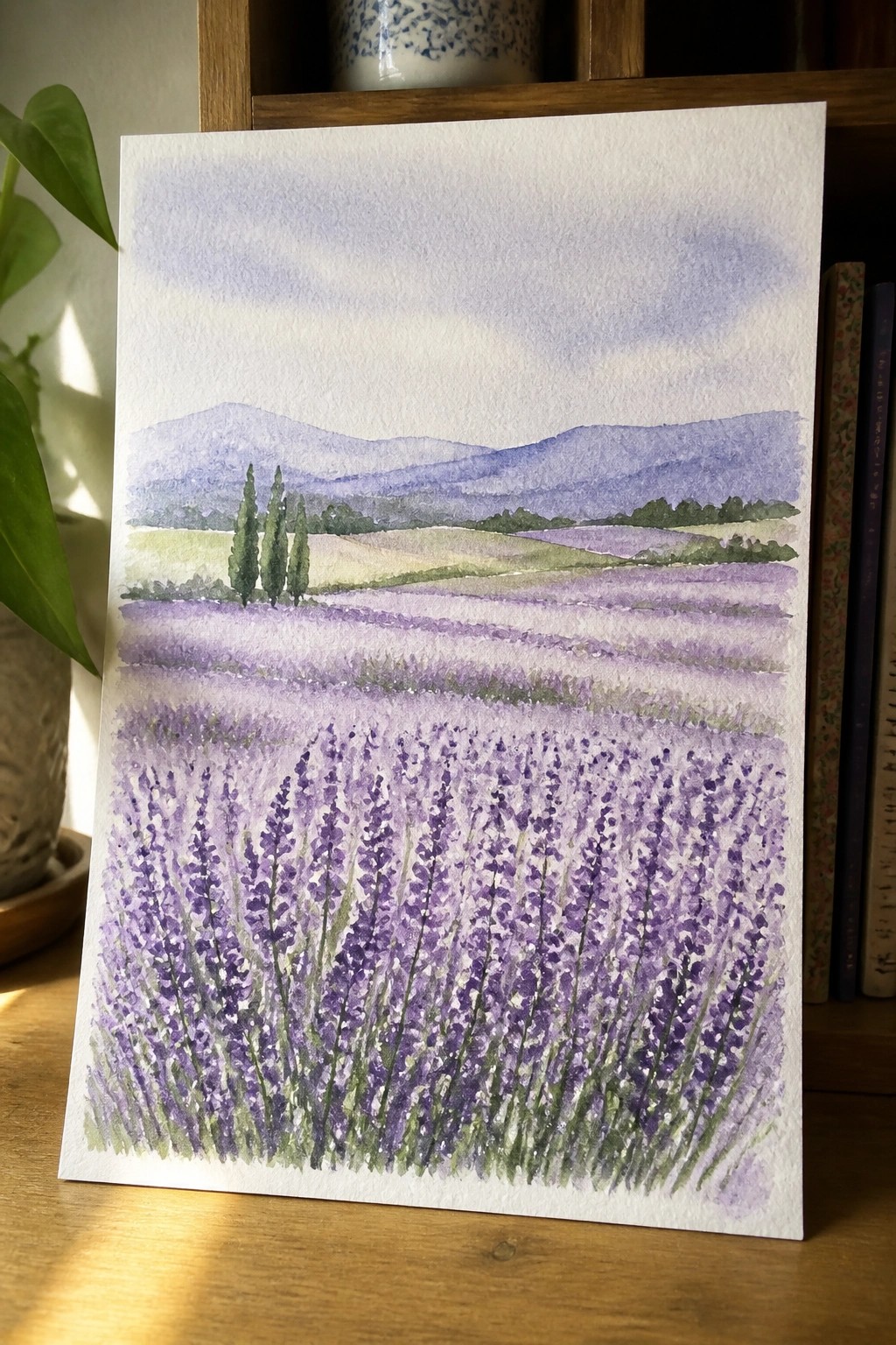 Watercolor painting of lavender fields with purple mountains on wooden table beside books