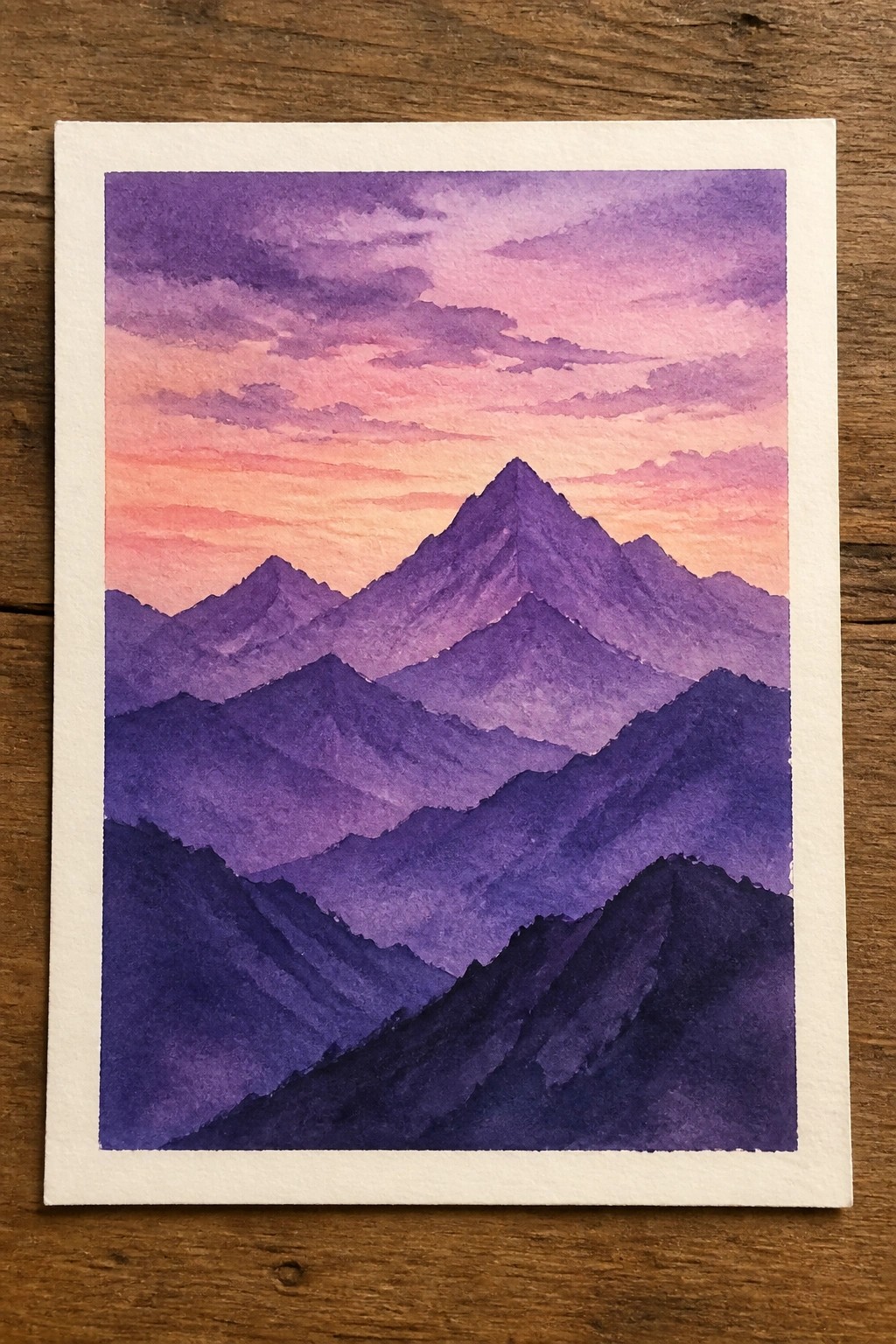 Watercolor painting of layered purple mountains beneath a pink-orange sunset sky.
