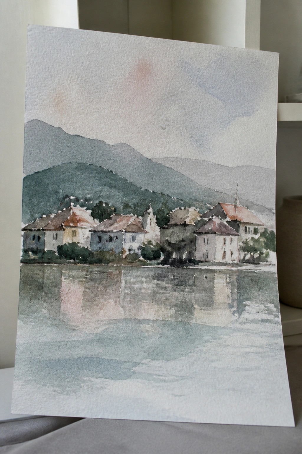Watercolor village with colorful houses reflected in a calm lake before green mountains.