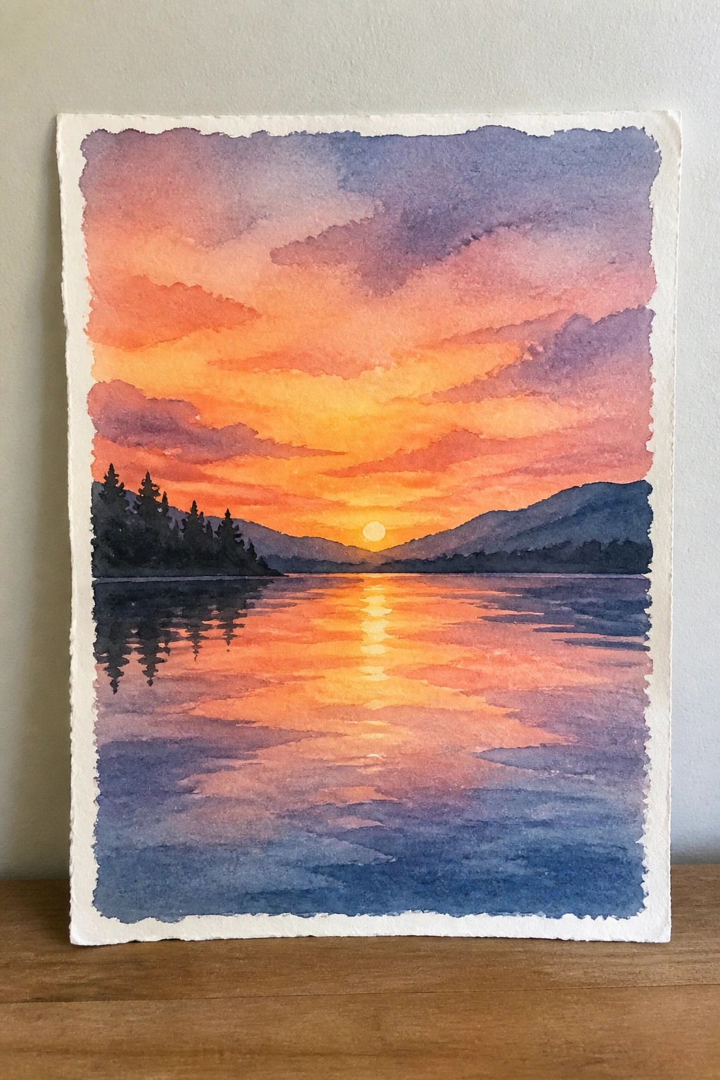 Watercolor sunset over lake with orange-pink sky, silhouetted mountains, trees, and reflections.