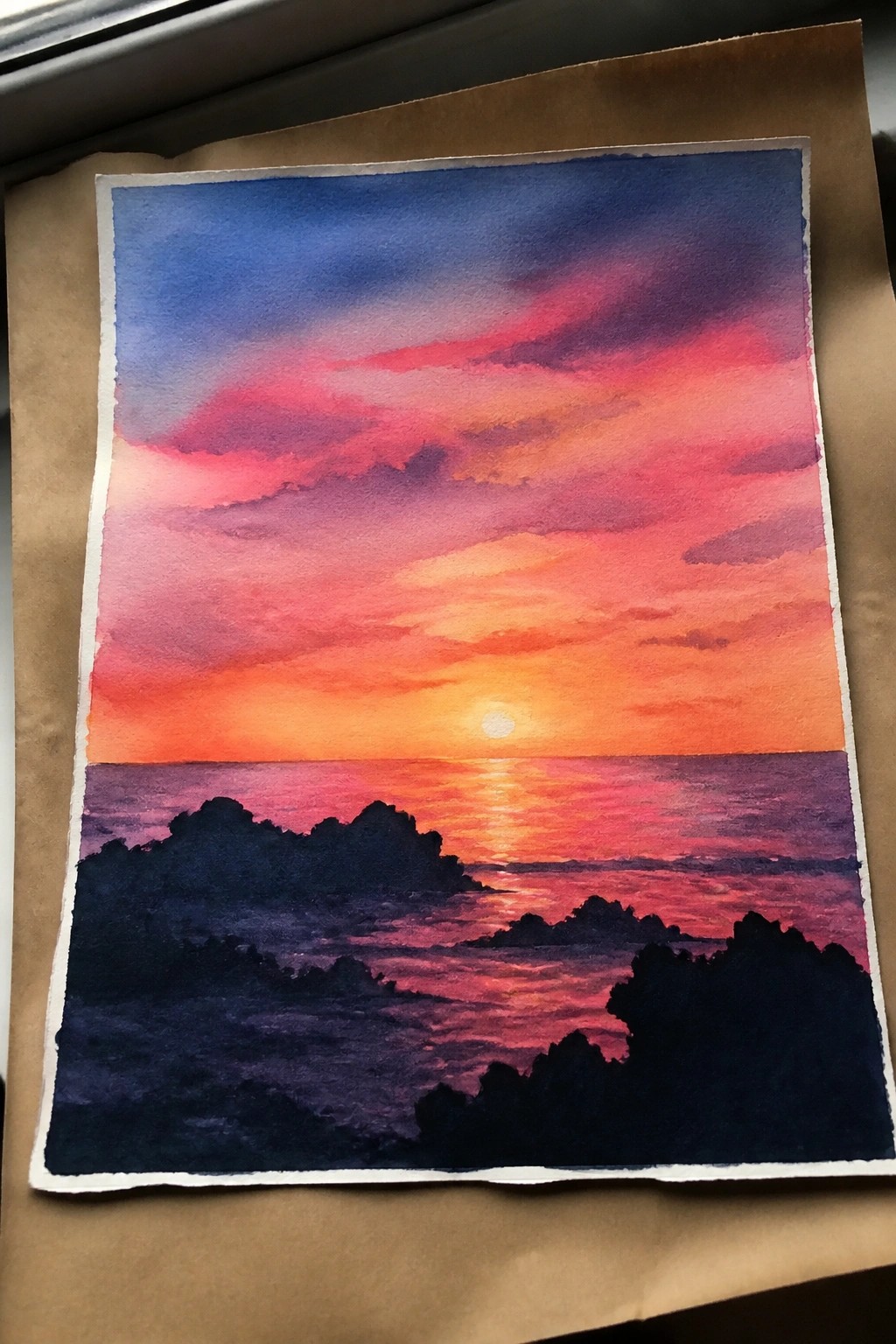 Vibrant watercolor sunset over ocean with pink-orange sky and dark rocky silhouettes.