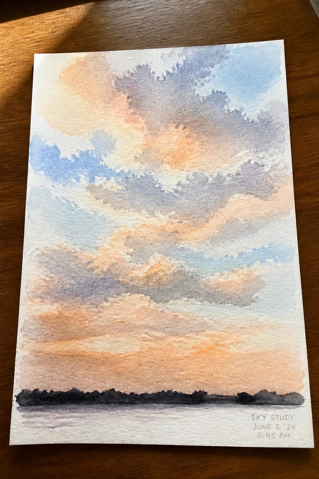 Watercolor painting of layered sunset clouds in orange, blue, and purple over dark horizon.