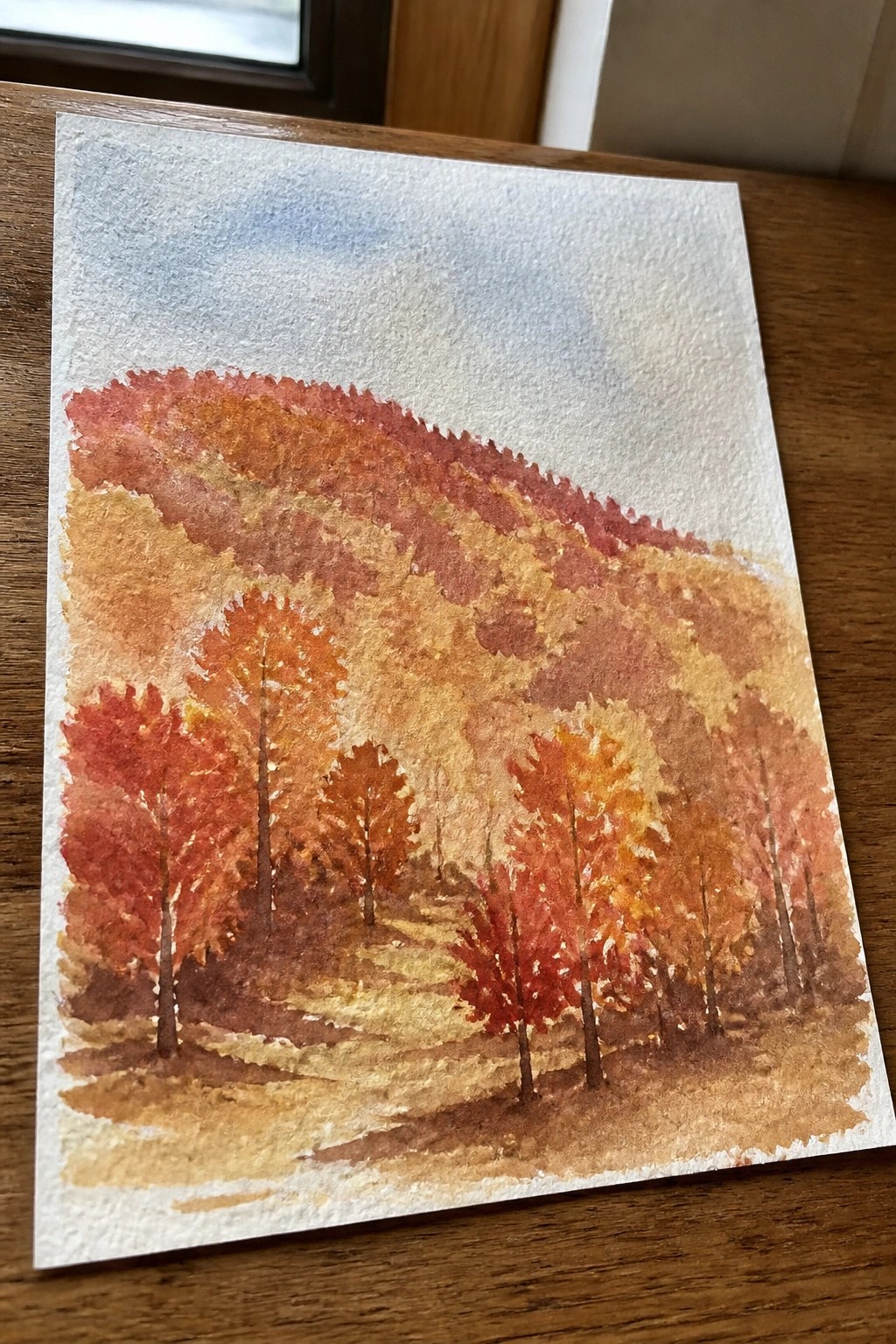 Watercolor painting of red and orange autumn trees on a forested hillside