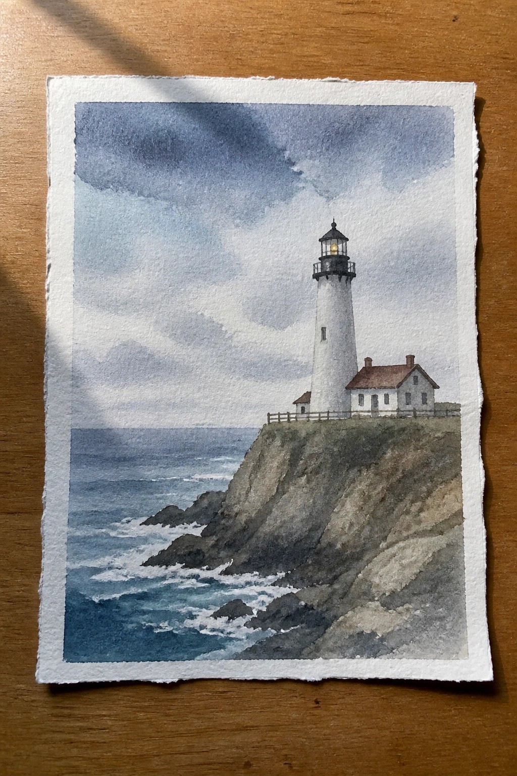 A watercolor painting of a white lighthouse with a red-roofed house on a rocky cliff above a blue ocean under cloudy skies.