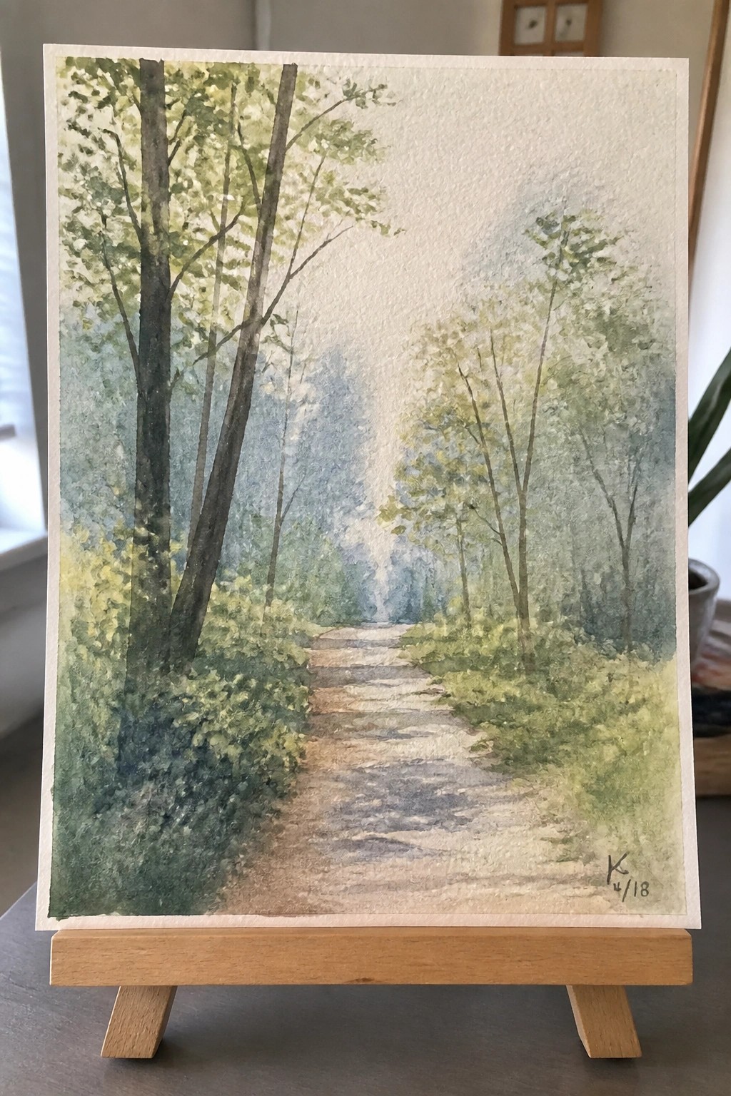 Watercolor of a misty dirt path winding through green trees on a wooden easel