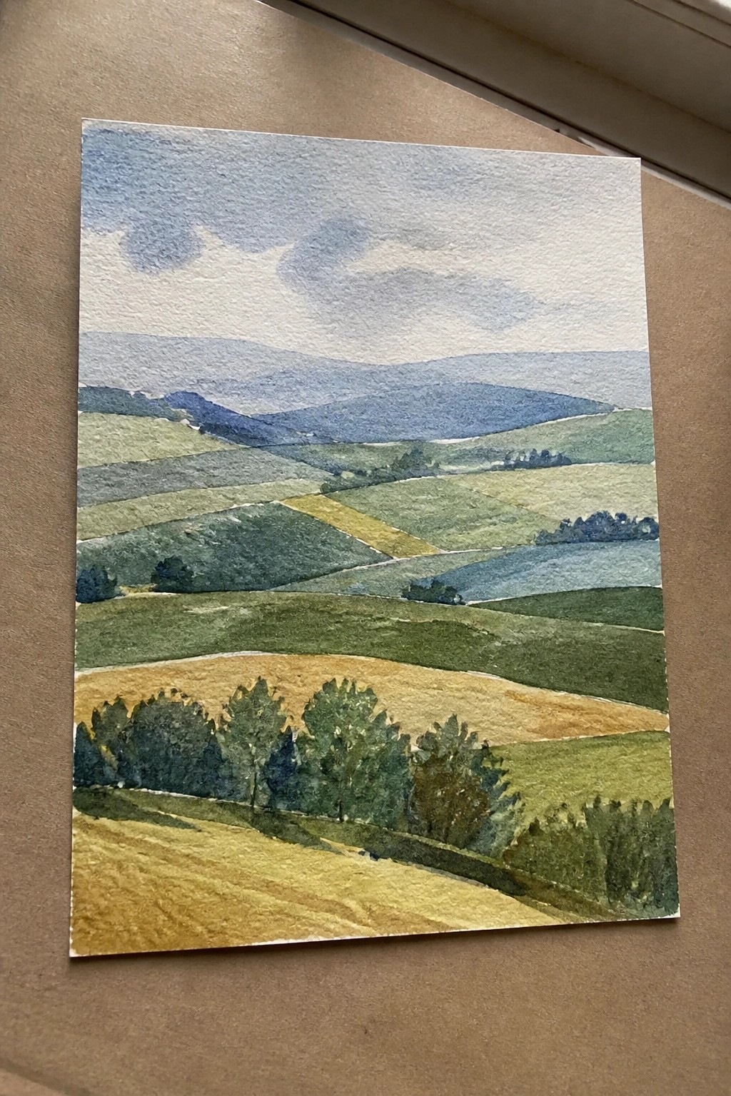 Watercolor landscape of rolling hills with patchwork fields in greens, yellows, and blues.