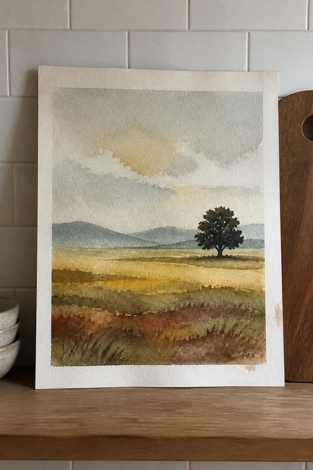 Watercolor landscape of golden fields with lone tree, distant blue mountains, and cloudy sky