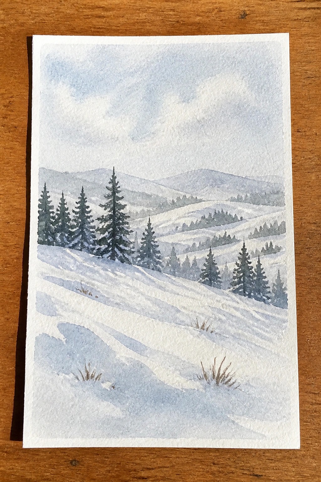 Watercolor of snow-covered hills with pine trees under a cloudy sky on wood