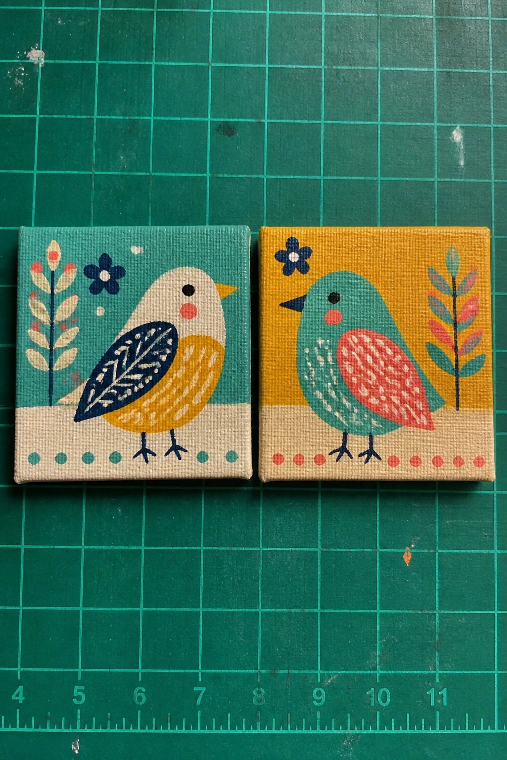 Two small square canvases with stylized birds on teal and yellow backgrounds sit on a green cutting mat.